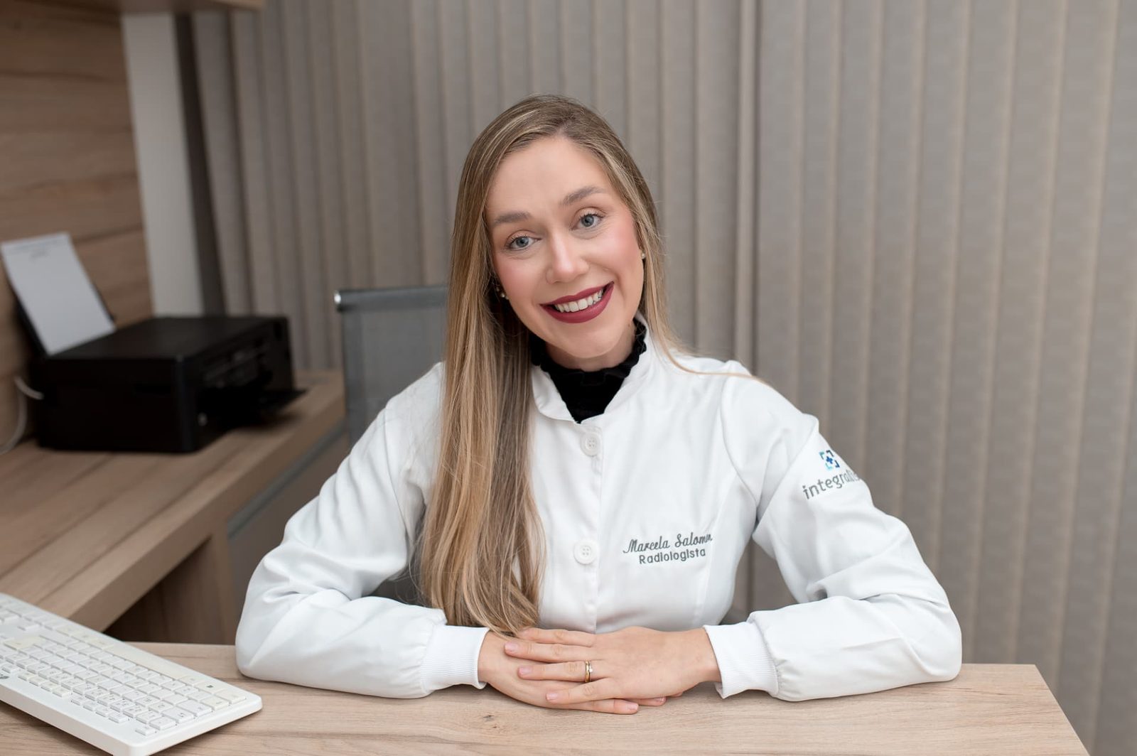 Dra. Marcela Salomon sentada à mesa do consultório, sorrindo durante o atendimento a um paciente